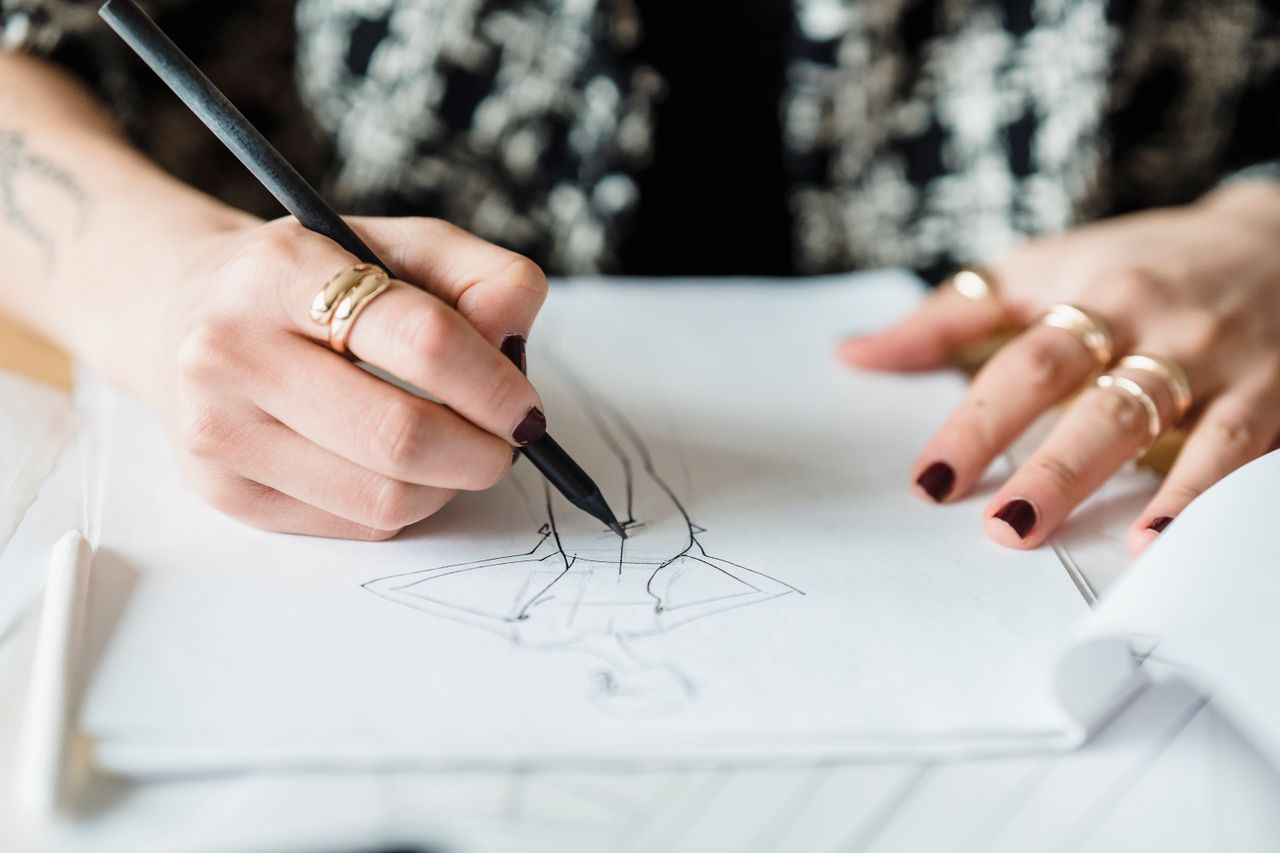 journey Crop unrecognizable female with rings drawing draft of modern garment while sitting at table with papers in light modern workshop